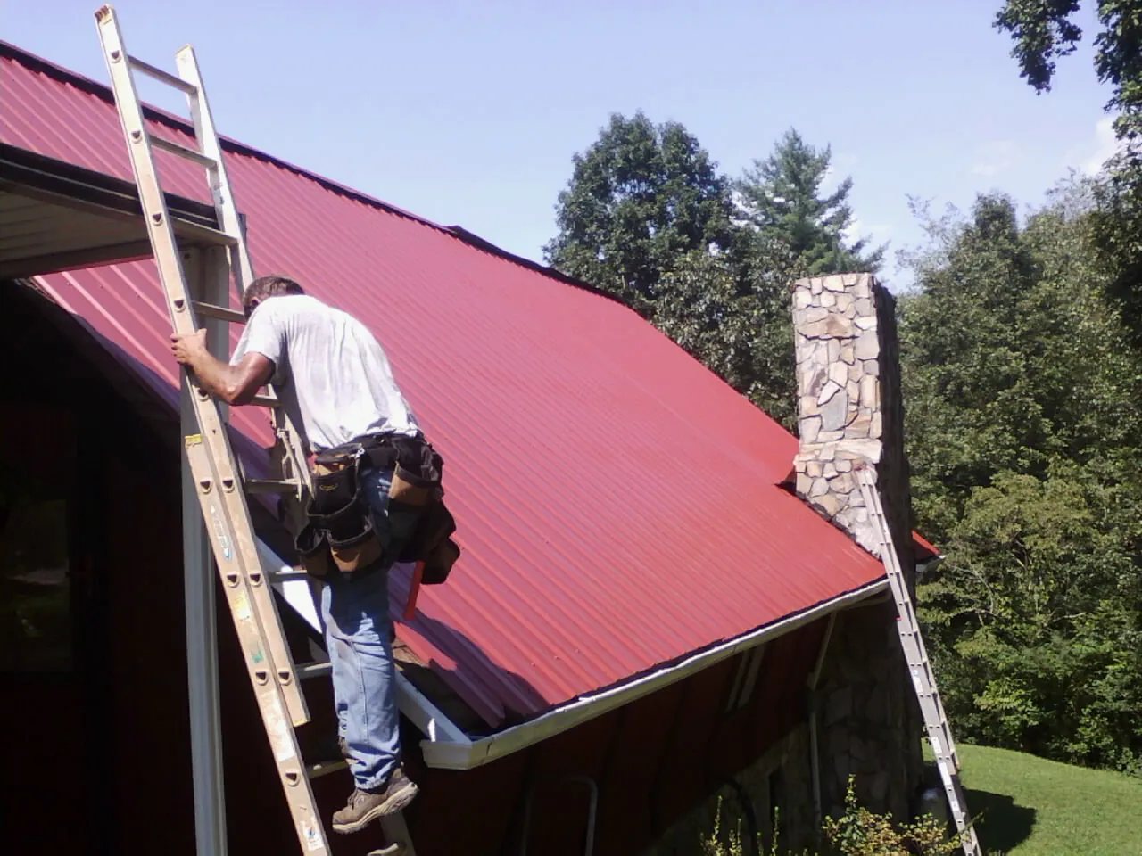Roofer on a ladder beside a red metal roof and stone chimney — Soffit & Fascia Repair by Roofing Brothers in Minneota